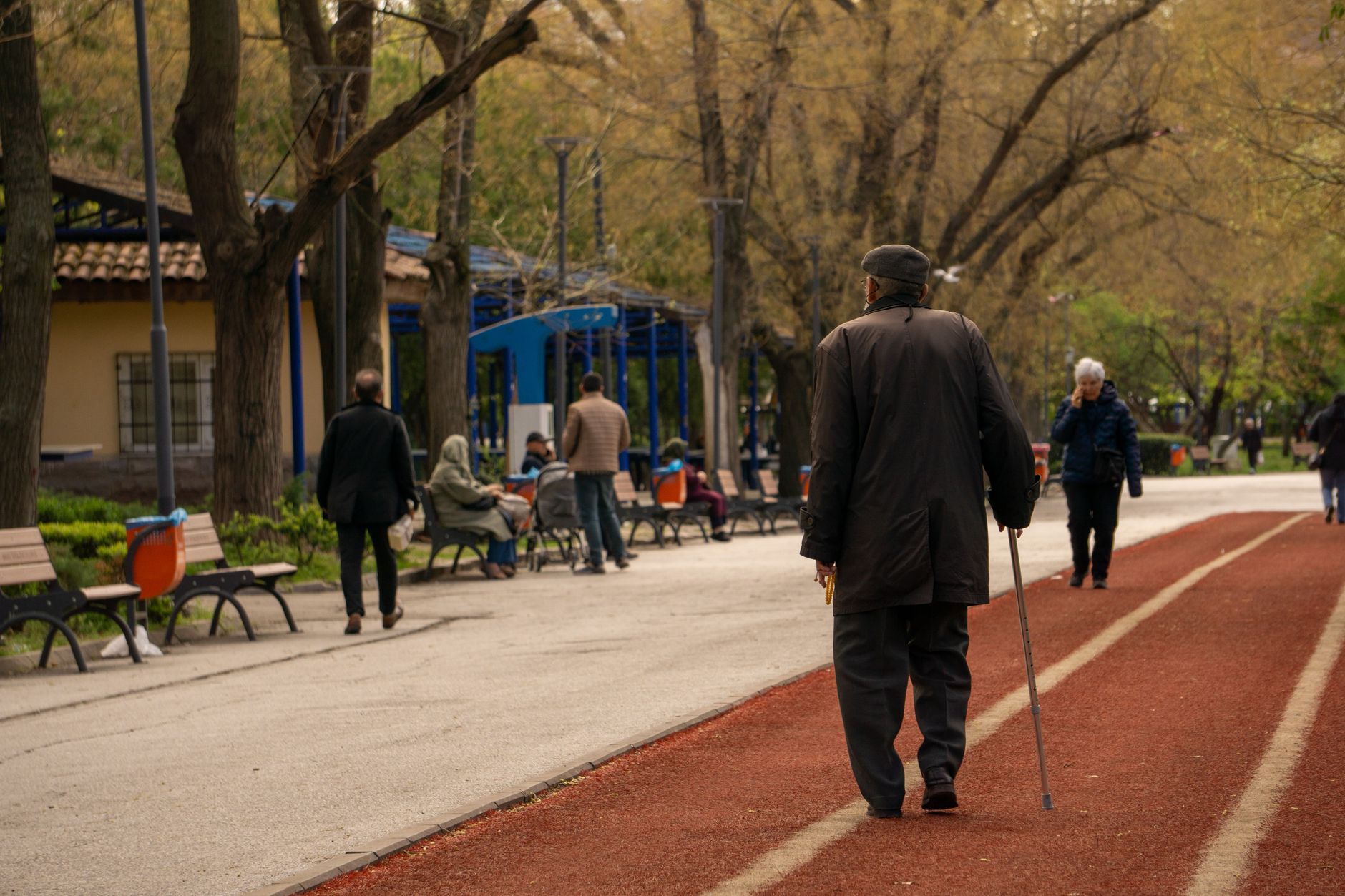 An elderly man with a cane strolls down a serene park path lined with benches and trees.